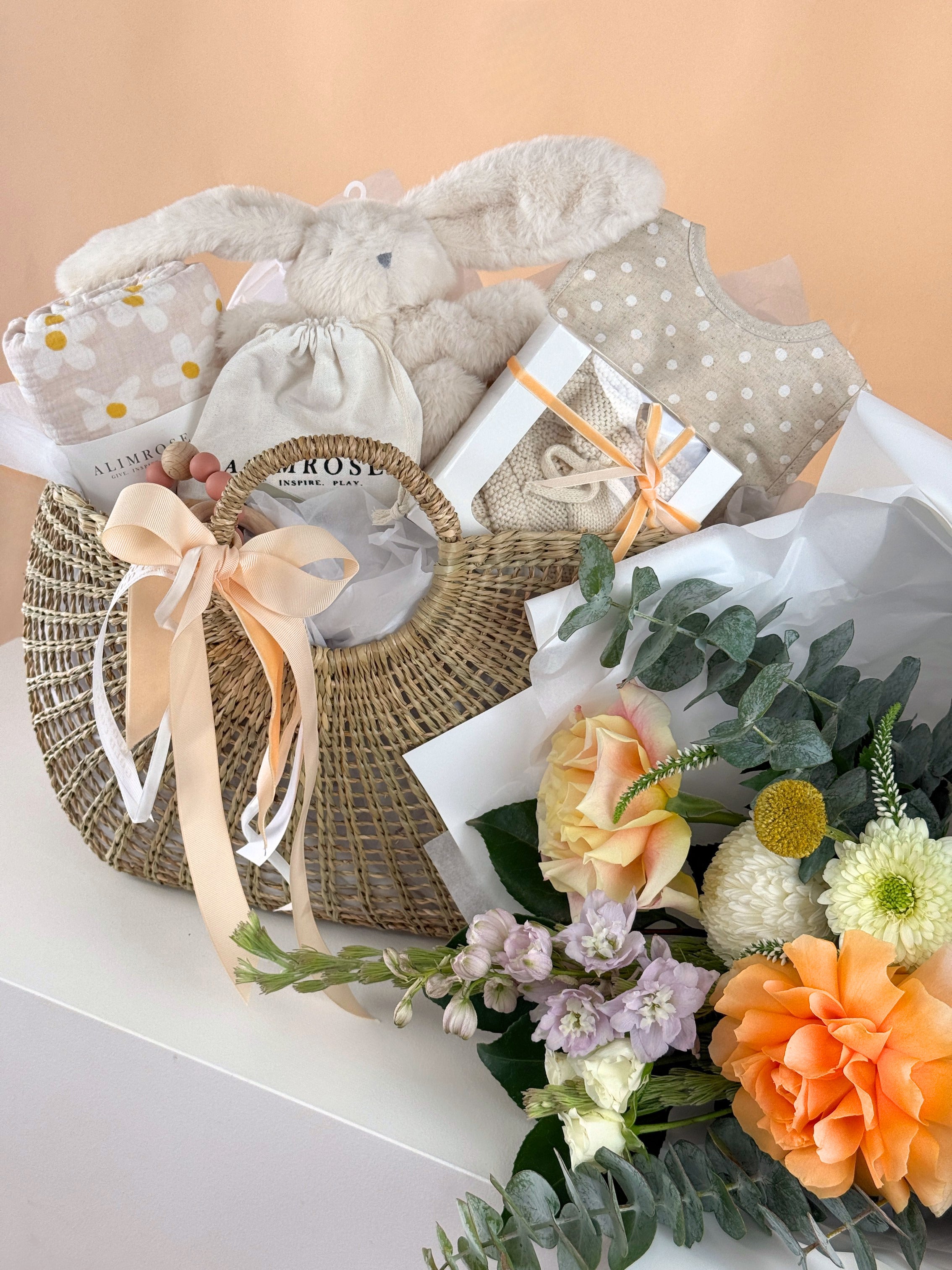 Baby gift basket with wicker bag, baby clothes, and flowers on a white surface, from Flowers Gold Coast